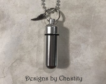 May include: A silver-colored cylindrical pendant on a ball chain, featuring a small metal wing charm attached to the jump ring. The pendant has a black rubber seal at the center. The text "Designs by Chastity" is printed below the object.