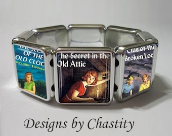 May include: Silver-toned stretch bracelet featuring Nancy Drew book covers. The bracelet showcases images from "The Secret of the Old Clock," "The Secret in the Old Attic," and "Clue of the Broken Lock." The text "Designs by Chastity" is at the bottom.