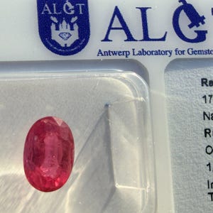Könnte beinhalten: Ein oval geschliffener, transparenter roter Rubin mit einem Gewicht von 1,31 Karat, eingeschlossen in einer durchsichtigen Kunststoffhülle. Der Edelstein wird von einem Bericht des ALGT Antwerp Laboratory for Gemstone Testing begleitet.