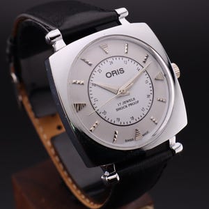 Puede incluir: Un reloj Oris plateado con una correa de cuero negro. La esfera del reloj es blanca con agujas y marcadores plateados. El reloj indica "17 Jewels Shock Proof" y "Swiss Made".