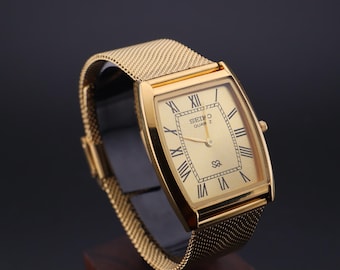 May include: A gold-toned Seiko Quartz watch with a rectangular face and mesh band. The watch face features black Roman numerals and hands, with the Seiko Quartz logo. The watch is displayed against a dark background.