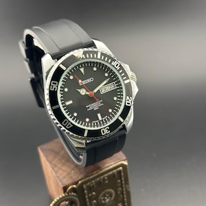 Pode incluir: Um relógio Seiko Marinemaster Professional 300m com mostrador preto, ponteiros prateados e um ponteiro de segundos vermelho. O relógio possui uma luneta giratória preta com números brancos, uma janela de dia e data às três horas e uma pulseira preta.