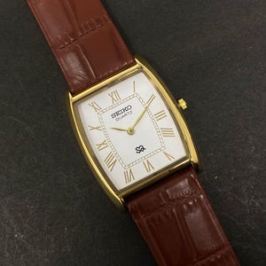 May include: A gold-toned Seiko Quartz watch with a rectangular face and brown leather strap. The white watch face features gold Roman numerals and hands. The brown leather strap has a textured, crocodile-like pattern.
