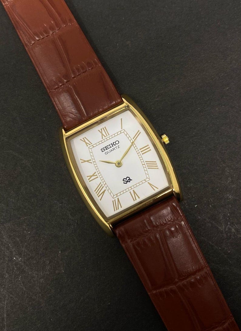 May include: A gold-toned Seiko Quartz watch with a rectangular face and brown leather strap. The white watch face features gold Roman numerals and hands. The brown leather strap has a textured, crocodile-like pattern.