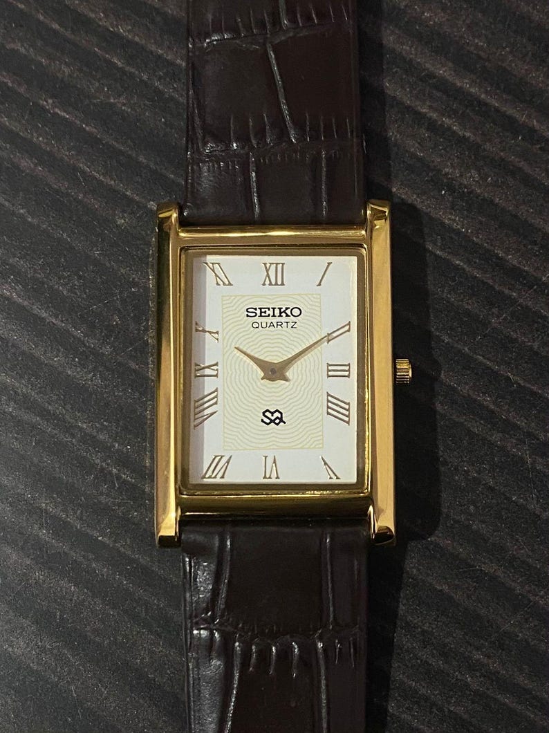 May include: A gold-toned Seiko Quartz watch with a rectangular face and brown leather band. The watch face is white with Roman numerals and gold hands. The band is dark brown with a textured pattern.