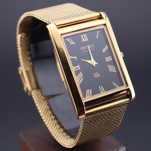 Könnte beinhalten: Eine goldfarbene Seiko Quartz-Uhr mit einem rechteckigen schwarzen Zifferblatt und goldenen römischen Ziffern. Die Uhr hat ein Mesh-Armband und wird vor einem dunklen Hintergrund präsentiert. Das Zifferblatt zeigt das Seiko Quartz-Logo.