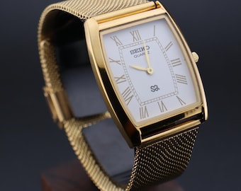 May include: A gold-toned Seiko Quartz watch with a rectangular face and mesh band. The watch face is white with gold Roman numerals and hands. The watch is displayed on a dark surface.