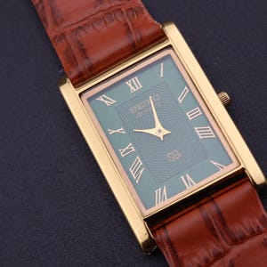 K&ouml;nnte beinhalten: Eine rechteckige, goldfarbene Armbanduhr mit einem t&uuml;rkisgr&uuml;nen Zifferblatt und r&ouml;mischen Ziffern. Die Uhr hat goldene Zeiger und ein braunes Lederarmband mit Krokodilmuster. Auf dem Zifferblatt steht die Marke "SEIKO".