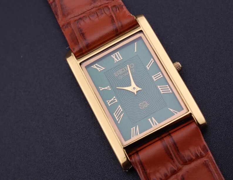 K&ouml;nnte beinhalten: Eine rechteckige, goldfarbene Armbanduhr mit einem t&uuml;rkisgr&uuml;nen Zifferblatt und r&ouml;mischen Ziffern. Die Uhr hat goldene Zeiger und ein braunes Lederarmband mit Krokodilmuster. Auf dem Zifferblatt steht die Marke "SEIKO".