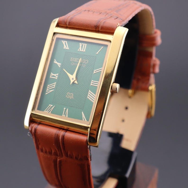 K&ouml;nnte beinhalten: Eine goldfarbene rechteckige Seiko Quartz-Uhr mit gr&uuml;nem Zifferblatt und r&ouml;mischen Ziffern. Die Uhr hat ein braunes Lederarmband mit Krokodilmuster. Das Zifferblatt zeigt das Seiko Quartz-Logo.