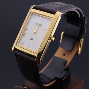 以下が含まれることがあります： 白い文字盤、ローマ数字、茶色の革バンドを備えた、金色の長方形のセイコー クォーツ腕時計。 金色のベゼルとテクスチャ加工された白い文字盤が特徴です。 バンドは濃い茶色で、バックル留めです。