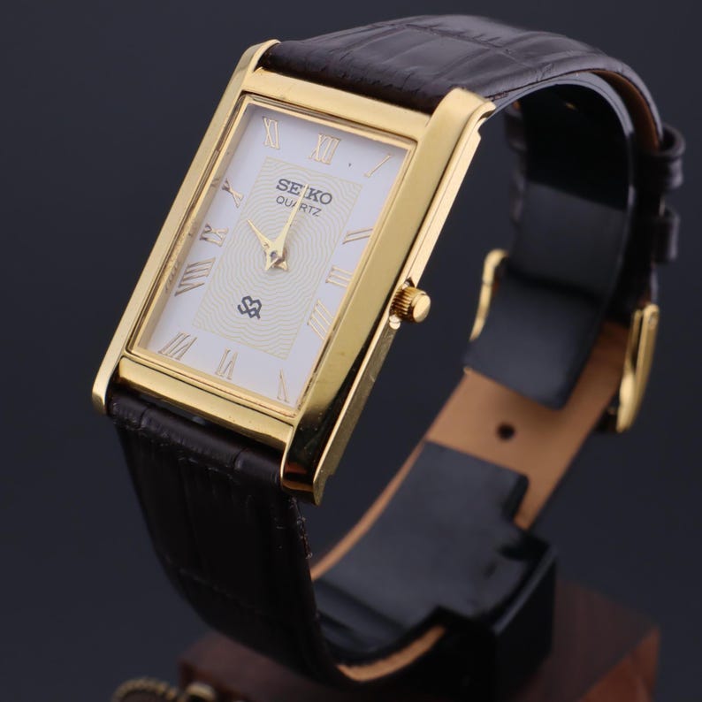 以下が含まれることがあります： 白い文字盤、ローマ数字、茶色の革バンドを備えた、金色の長方形のセイコー クォーツ腕時計。 金色のベゼルとテクスチャ加工された白い文字盤が特徴です。 バンドは濃い茶色で、バックル留めです。