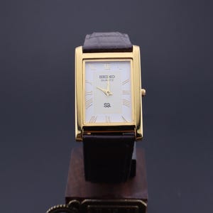 以下が含まれることがあります： 長方形の文字盤と濃い茶色の革バンドが付いた、金色のセイコー クォーツ腕時計。 文字盤は白で、金色の針とローマ数字の時字が付いています。 時計は木製のスタンドに展示されています。