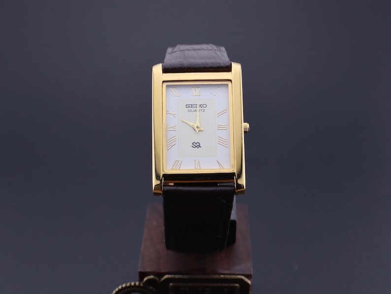 以下が含まれることがあります： 長方形の文字盤と濃い茶色の革バンドが付いた、金色のセイコー クォーツ腕時計。 文字盤は白で、金色の針とローマ数字の時字が付いています。 時計は木製のスタンドに展示されています。