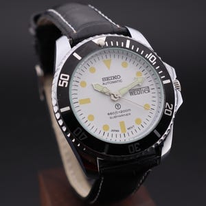 Peut inclure: Montre Seiko automatique Submariner avec lunette noire et cadran blanc. La montre est dotée d'un bracelet en cuir noir et d'un affichage de la date. Le cadran comporte des marqueurs et des aiguilles luminescents. Résistant à l'eau jusqu'à 200m.