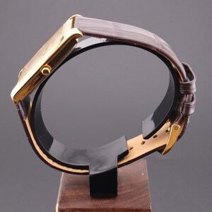 以下が含まれることがあります： 茶色の革バンドが付いた、金色の長方形の腕時計。 時計は、時計とバンドの側面図を示す、木製のベースが付いた黒いスタンドに展示されています。 時計はクラシックでエレガントなデザインです。