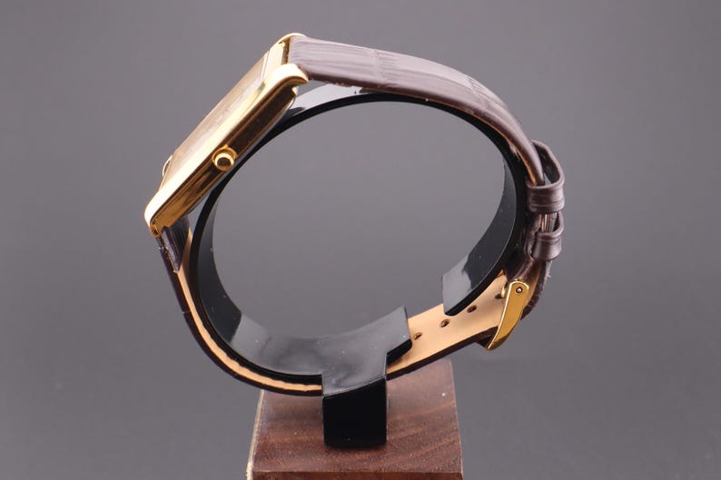 以下が含まれることがあります： 茶色の革バンドが付いた、金色の長方形の腕時計。 時計は、時計とバンドの側面図を示す、木製のベースが付いた黒いスタンドに展示されています。 時計はクラシックでエレガントなデザインです。