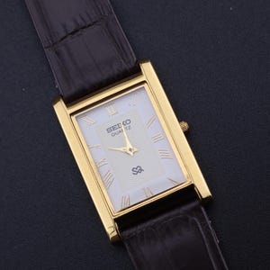 以下が含まれることがあります： 長方形の金色のセイコー クォーツ腕時計。白い文字盤にローマ数字のインデックス。黒い革バンドが付いています。文字盤にはセイコーのロゴと「Quartz」の文字が表示されています。