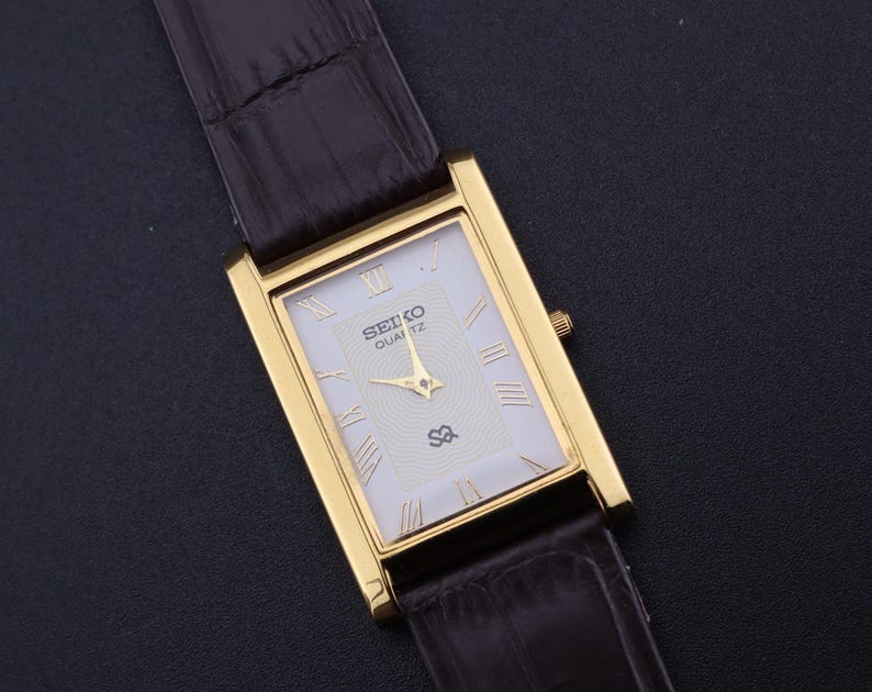 以下が含まれることがあります： 長方形の金色のセイコー クォーツ腕時計。白い文字盤にローマ数字のインデックス。黒い革バンドが付いています。文字盤にはセイコーのロゴと「Quartz」の文字が表示されています。