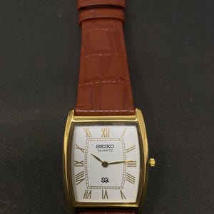 May include: A gold-toned Seiko Quartz watch with a rectangular face and brown leather strap. The white watch face features gold Roman numerals and hands. The strap is a rich brown colour with a textured pattern.