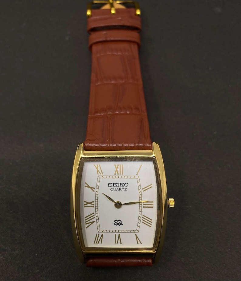 May include: A gold-toned Seiko Quartz watch with a rectangular face and brown leather strap. The white watch face features gold Roman numerals and hands. The strap is a rich brown colour with a textured pattern.