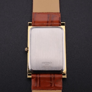 K&ouml;nnte beinhalten: Eine goldfarbene rechteckige Uhr mit einem braunen Lederarmband. Das Zifferblatt der Uhr ist silberfarben und das Armband ist rotbraun. Die Uhr hat ein klassisches, Vintage-Design.