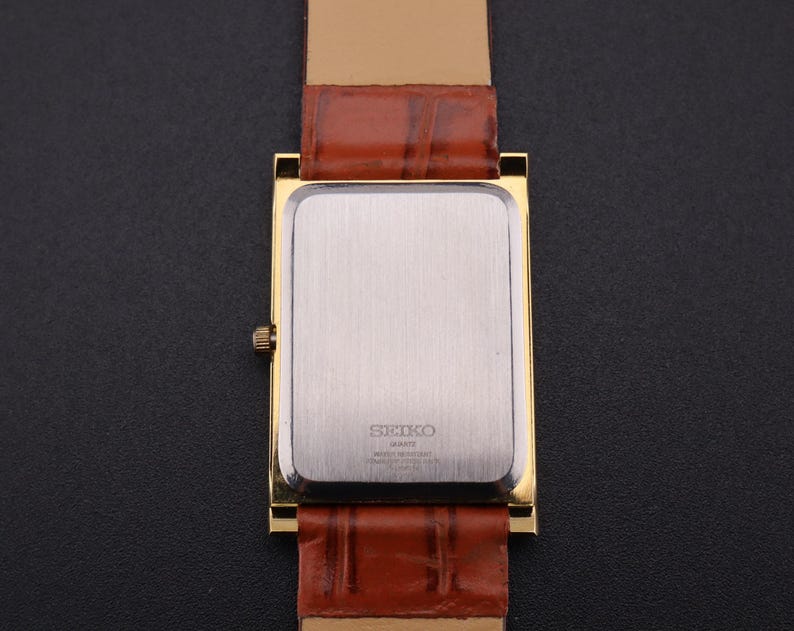K&ouml;nnte beinhalten: Eine goldfarbene rechteckige Uhr mit einem braunen Lederarmband. Das Zifferblatt der Uhr ist silberfarben und das Armband ist rotbraun. Die Uhr hat ein klassisches, Vintage-Design.