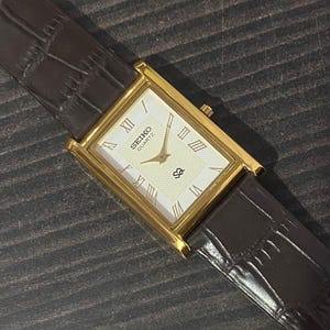 May include: A gold-toned Seiko Quartz watch with a rectangular face and a brown leather band. The watch face is cream-colored with Roman numeral hour markers and the Seiko logo. The band has a crocodile skin texture.