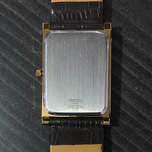 May include: A gold-toned Seiko quartz watch with a rectangular face and a dark brown leather strap. The watch back is silver with the Seiko logo and text. The strap is attached to the watch with gold-toned metal. The watch is on a dark surface.