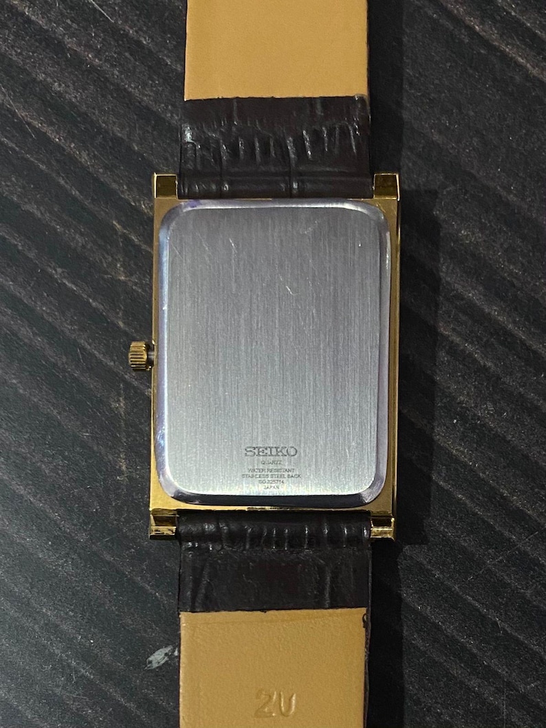 May include: A gold-toned Seiko quartz watch with a rectangular face and a dark brown leather strap. The watch back is silver with the Seiko logo and text. The strap is attached to the watch with gold-toned metal. The watch is on a dark surface.