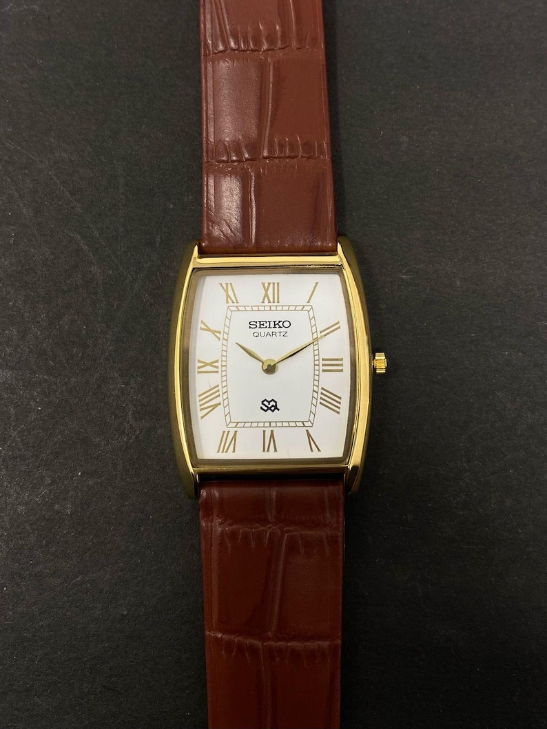 May include: A gold-coloured Seiko Quartz watch with a rectangular white face and Roman numeral hour markers. The watch has a brown leather strap with a crocodile pattern. The watch face displays the Seiko logo and the letters "SQ".