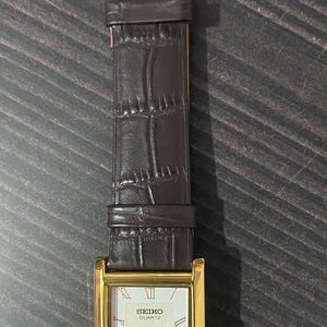 May include: A rectangular gold-toned Seiko Quartz watch with a brown leather band. The watch face is cream-colored with black Roman numerals and hands. The band has a crocodile pattern and a gold-colored buckle.