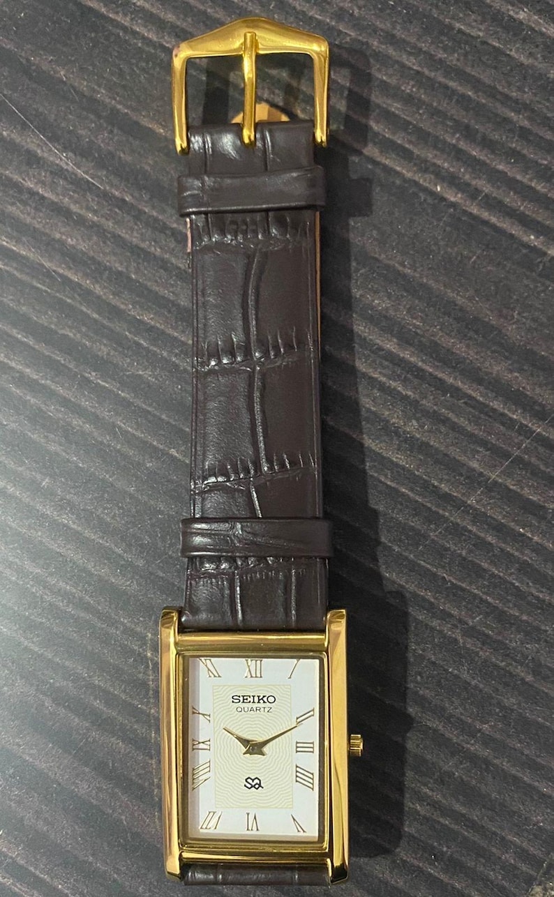 May include: A rectangular gold-toned Seiko Quartz watch with a brown leather band. The watch face is cream-colored with black Roman numerals and hands. The band has a crocodile pattern and a gold-colored buckle.
