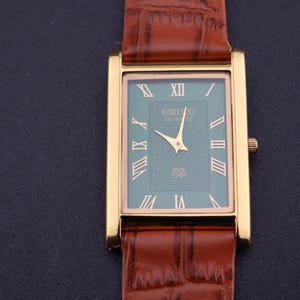 K&ouml;nnte beinhalten: Goldfarbene rechteckige Seiko Quartz-Uhr mit t&uuml;rkisgr&uuml;nem Zifferblatt und r&ouml;mischen Ziffern. Die Uhr hat goldene Zeiger und ein braunes Lederarmband mit Krokodilmuster.