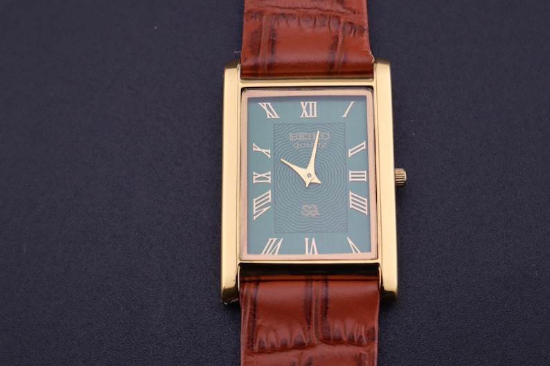 K&ouml;nnte beinhalten: Goldfarbene rechteckige Seiko Quartz-Uhr mit t&uuml;rkisgr&uuml;nem Zifferblatt und r&ouml;mischen Ziffern. Die Uhr hat goldene Zeiger und ein braunes Lederarmband mit Krokodilmuster.
