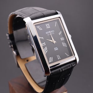 Peut inclure: Une montre Seiko Quartz rectangulaire avec un cadran noir et un boîtier argenté. La montre présente des chiffres romains et un bracelet en cuir noir. La montre est présentée sur un support en bois.