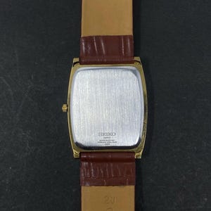 May include: A gold-plated Seiko quartz watch with a rectangular face and a brown leather strap. The watch has a silver-coloured back and a gold crown. The strap is a lighter brown on the inside and a darker brown on the outside.
