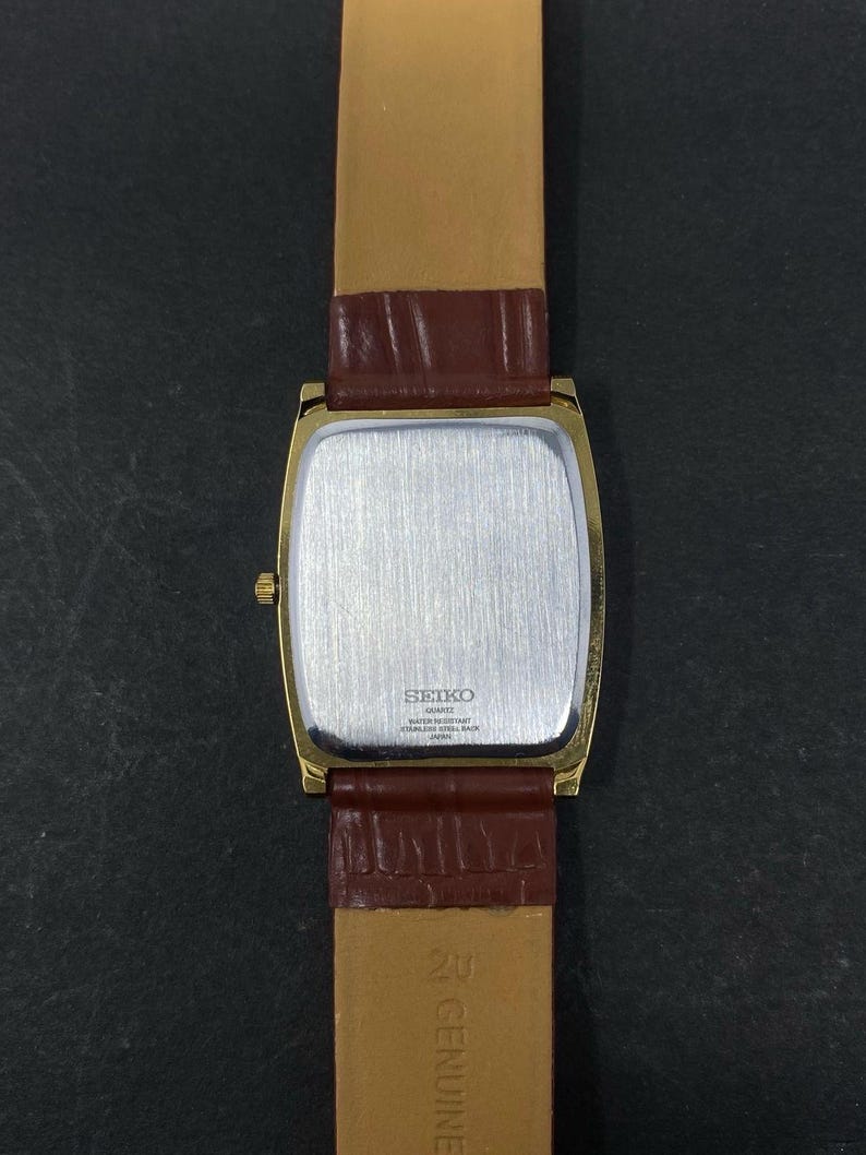 May include: A gold-plated Seiko quartz watch with a rectangular face and a brown leather strap. The watch has a silver-coloured back and a gold crown. The strap is a lighter brown on the inside and a darker brown on the outside.