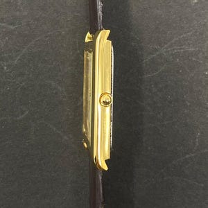 May include: A gold-toned rectangular wristwatch with a dark brown leather band. The watch face is white. The watch has a classic design. The band is thin and appears to be made of leather. The watch is viewed from the side.