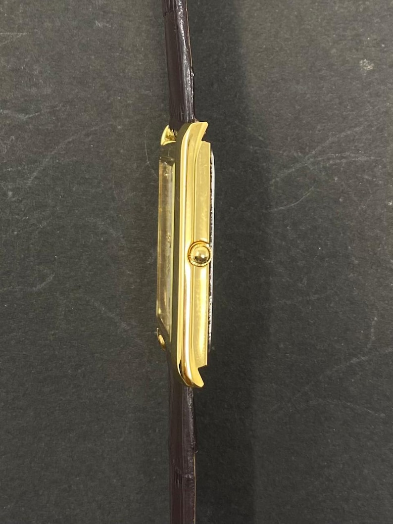 May include: A gold-toned rectangular wristwatch with a dark brown leather band. The watch face is white. The watch has a classic design. The band is thin and appears to be made of leather. The watch is viewed from the side.