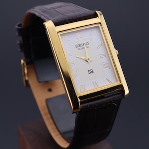 以下が含まれることがあります： 長方形の文字盤と黒い革バンドが付いた、金色のセイコー クォーツ腕時計。 文字盤は白で、金色のローマ数字と針が付いています。 時計は木製のスタンドに展示されています。