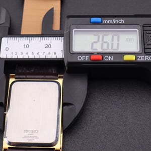 以下が含まれることがあります： 茶色の革バンドが付いた金色のセイコー腕時計をデジタルノギスで測定しています。時計の文字盤は長方形です。ノギスは26.0mmを示しています。
