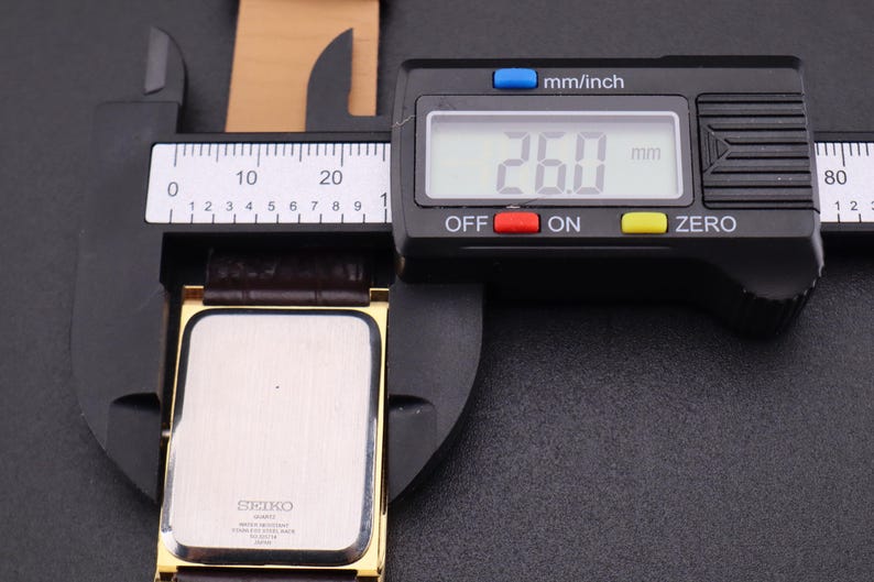 以下が含まれることがあります： 茶色の革バンドが付いた金色のセイコー腕時計をデジタルノギスで測定しています。時計の文字盤は長方形です。ノギスは26.0mmを示しています。