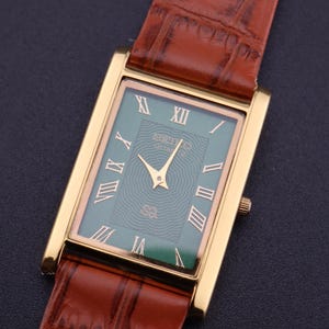 K&ouml;nnte beinhalten: Eine rechteckige, goldfarbene Armbanduhr mit gr&uuml;nem Zifferblatt und r&ouml;mischen Ziffern. Die Uhr hat goldene Zeiger und ein braunes Lederarmband. Auf dem Zifferblatt steht der Markenname "SEIKO". Die Uhr ist vor einem dunklen Hintergrund platziert.