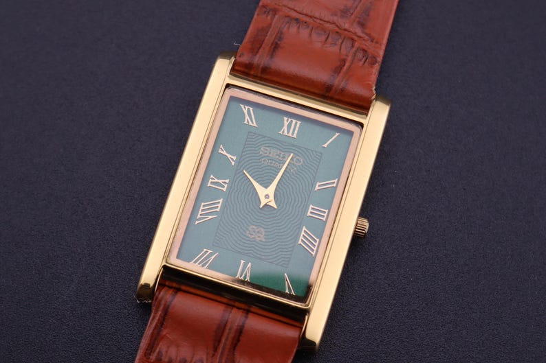 K&ouml;nnte beinhalten: Eine rechteckige, goldfarbene Armbanduhr mit gr&uuml;nem Zifferblatt und r&ouml;mischen Ziffern. Die Uhr hat goldene Zeiger und ein braunes Lederarmband. Auf dem Zifferblatt steht der Markenname "SEIKO". Die Uhr ist vor einem dunklen Hintergrund platziert.
