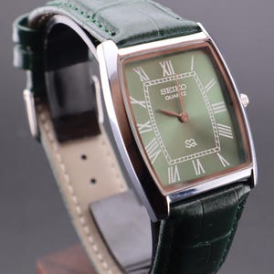 May include: A rectangular Seiko Quartz watch with a green face and Roman numeral hour markers. The watch has a silver case and a green leather strap. The watch face displays the Seiko Quartz logo and a small symbol.