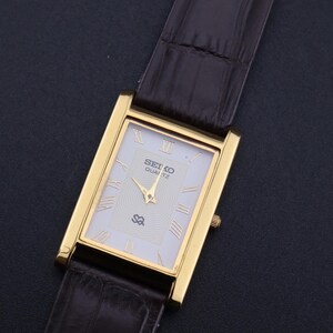 以下が含まれることがあります： 白い文字盤とローマ数字のインデックスを備えた、長方形のゴールドトーンのセイコー クォーツ腕時計。 金色の針と茶色の革バンドが付いています。 文字盤にはセイコー クォーツのロゴが表示されています。