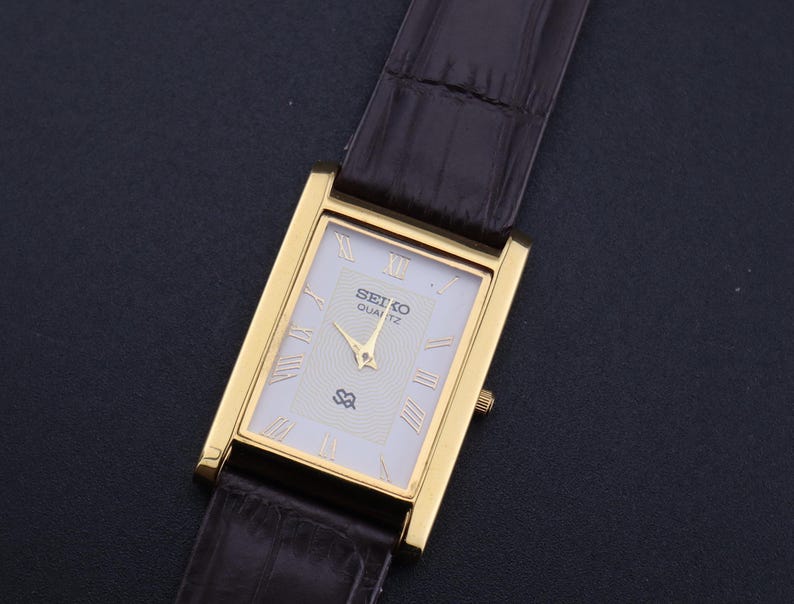 以下が含まれることがあります： 白い文字盤とローマ数字のインデックスを備えた、長方形のゴールドトーンのセイコー クォーツ腕時計。 金色の針と茶色の革バンドが付いています。 文字盤にはセイコー クォーツのロゴが表示されています。