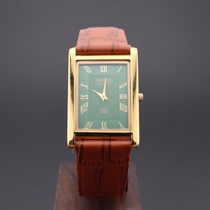 K&ouml;nnte beinhalten: Eine rechteckige Seiko-Uhr in Goldoptik mit gr&uuml;nem Zifferblatt und r&ouml;mischen Ziffern. Die Uhr hat goldene Zeiger und ein braunes Lederarmband. Die Uhr wird auf einem Holzst&auml;nder pr&auml;sentiert.