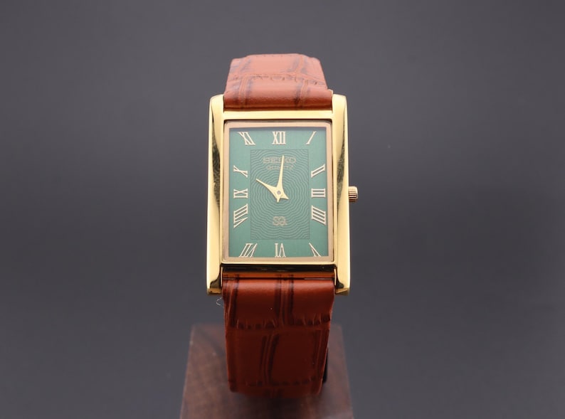 K&ouml;nnte beinhalten: Eine rechteckige Seiko-Uhr in Goldoptik mit gr&uuml;nem Zifferblatt und r&ouml;mischen Ziffern. Die Uhr hat goldene Zeiger und ein braunes Lederarmband. Die Uhr wird auf einem Holzst&auml;nder pr&auml;sentiert.